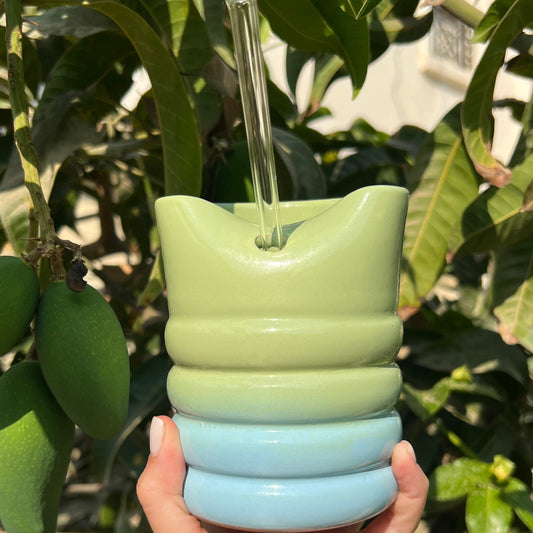 Hand holding a green and blue textured cup with a straw against a leafy background