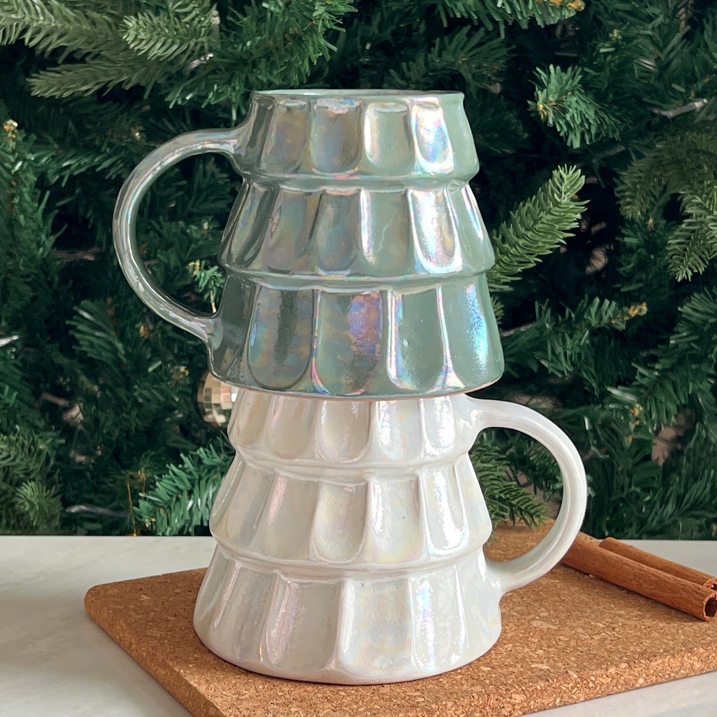 Two iridescent ceramic Christmas tree mugs, stacked, in festive green and white tones, placed in front of a decorated Christmas tree.